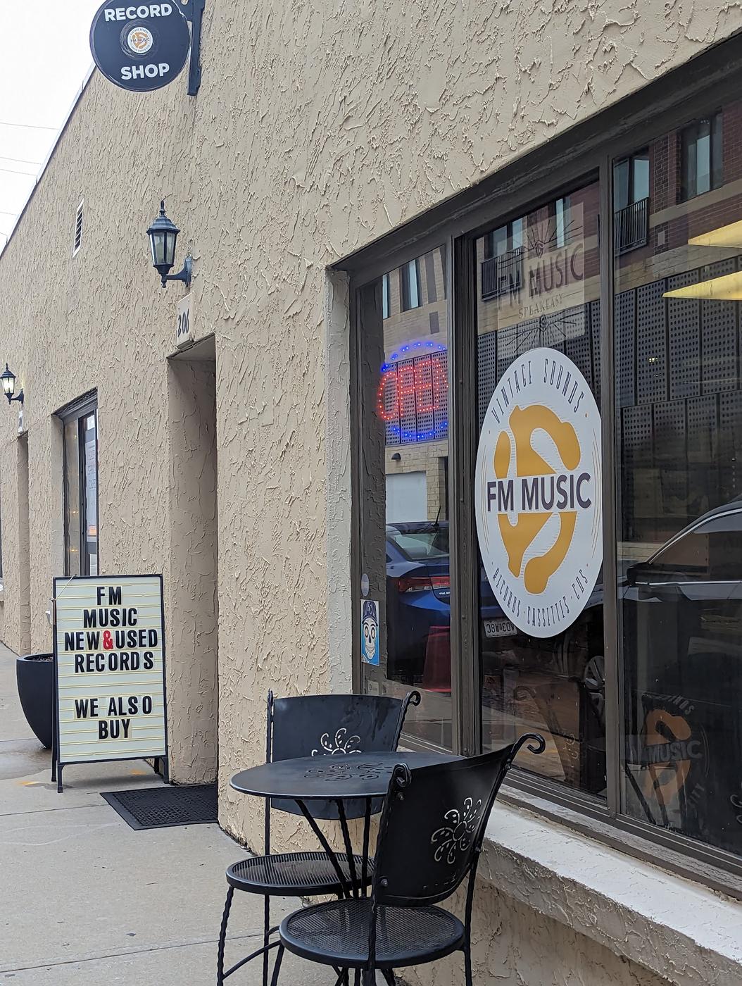 The FM Music storefront with its hanging record-shaped Record Shop sign, the window roundel, and an A-frame reading New and Used Records, We Also Buy.